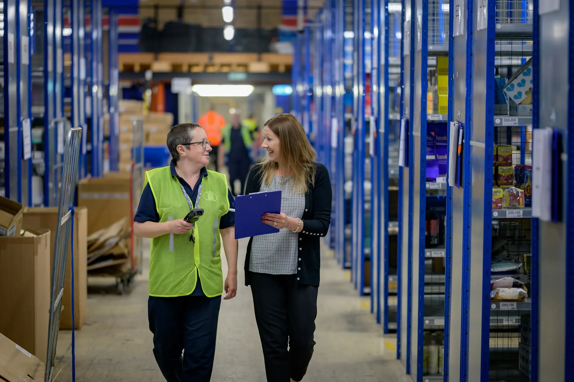 Two colleagues walking a warehouse aisle with a clipboard, between tall blue shelving.